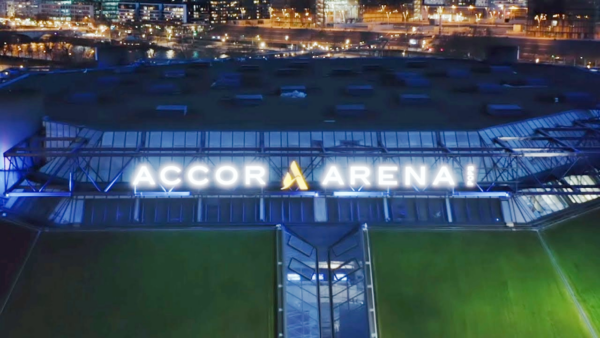 Veduta aerea notturna dell'esterno dell'Accor Arena con l'insegna illuminata e le luci della città sullo sfondo.