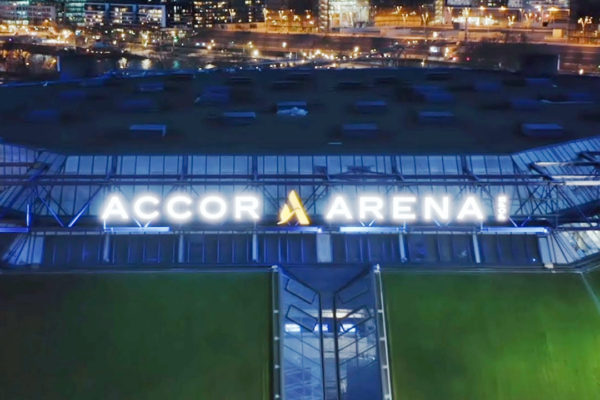 Veduta aerea notturna dell'esterno dell'Accor Arena con l'insegna illuminata e le luci della città sullo sfondo.