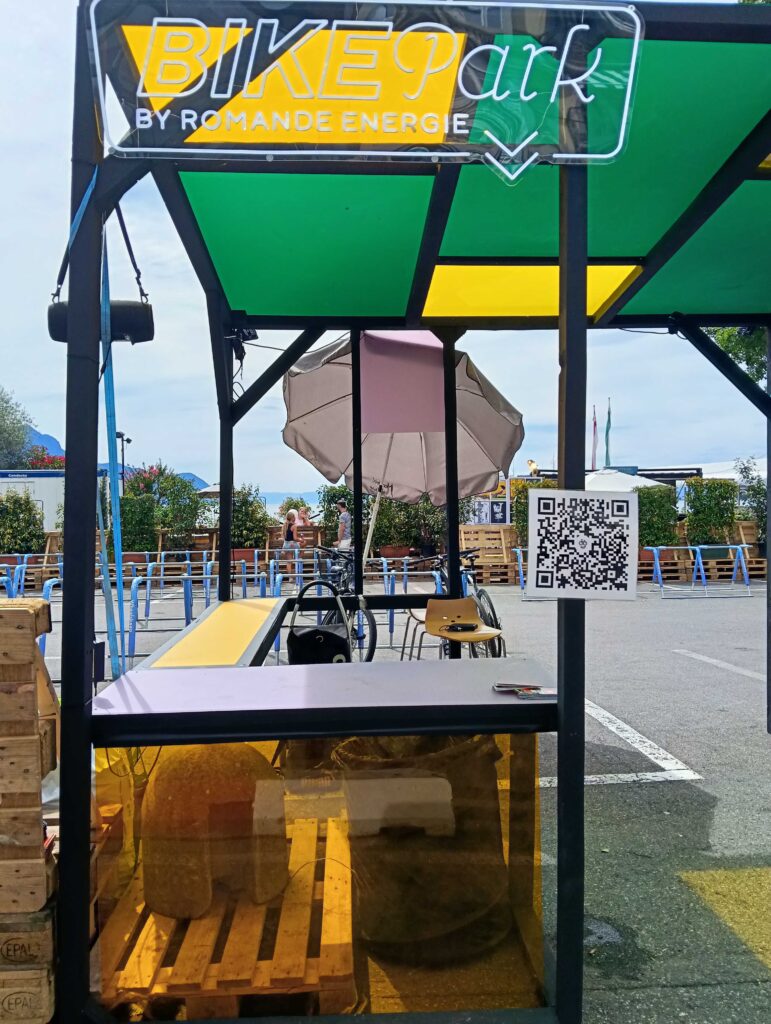 Chiosco per parcheggio bici all'aperto con insegna al neon “BIKEpark”, pannelli verdi e gialli, pallet in legno e un cartello con codice QR nelle vicinanze; persone e bici visibili sullo sfondo.