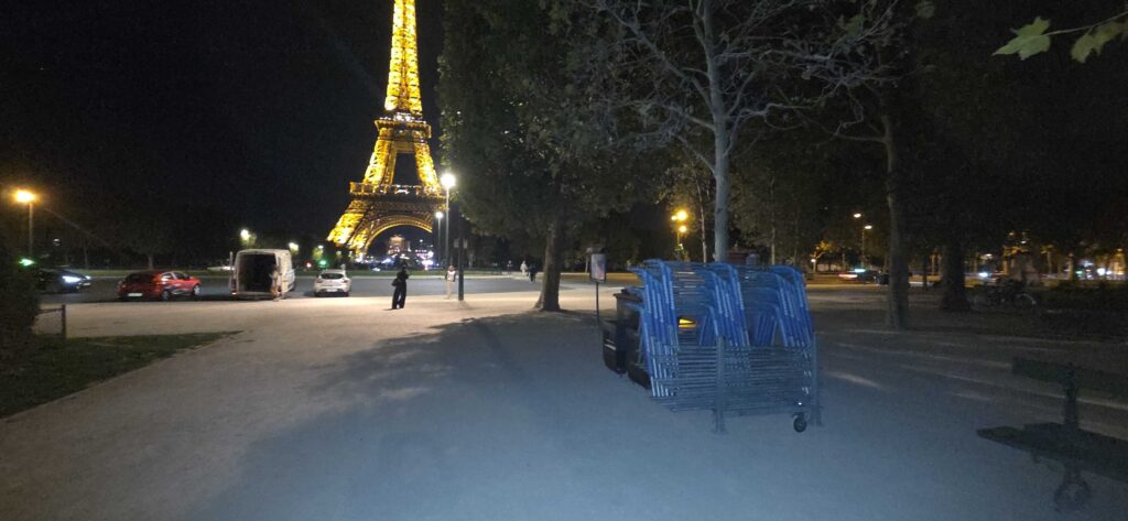 Vista notturna della Torre Eiffel illuminata di giallo, vista da un parco con sedie blu impilate in primo piano.
